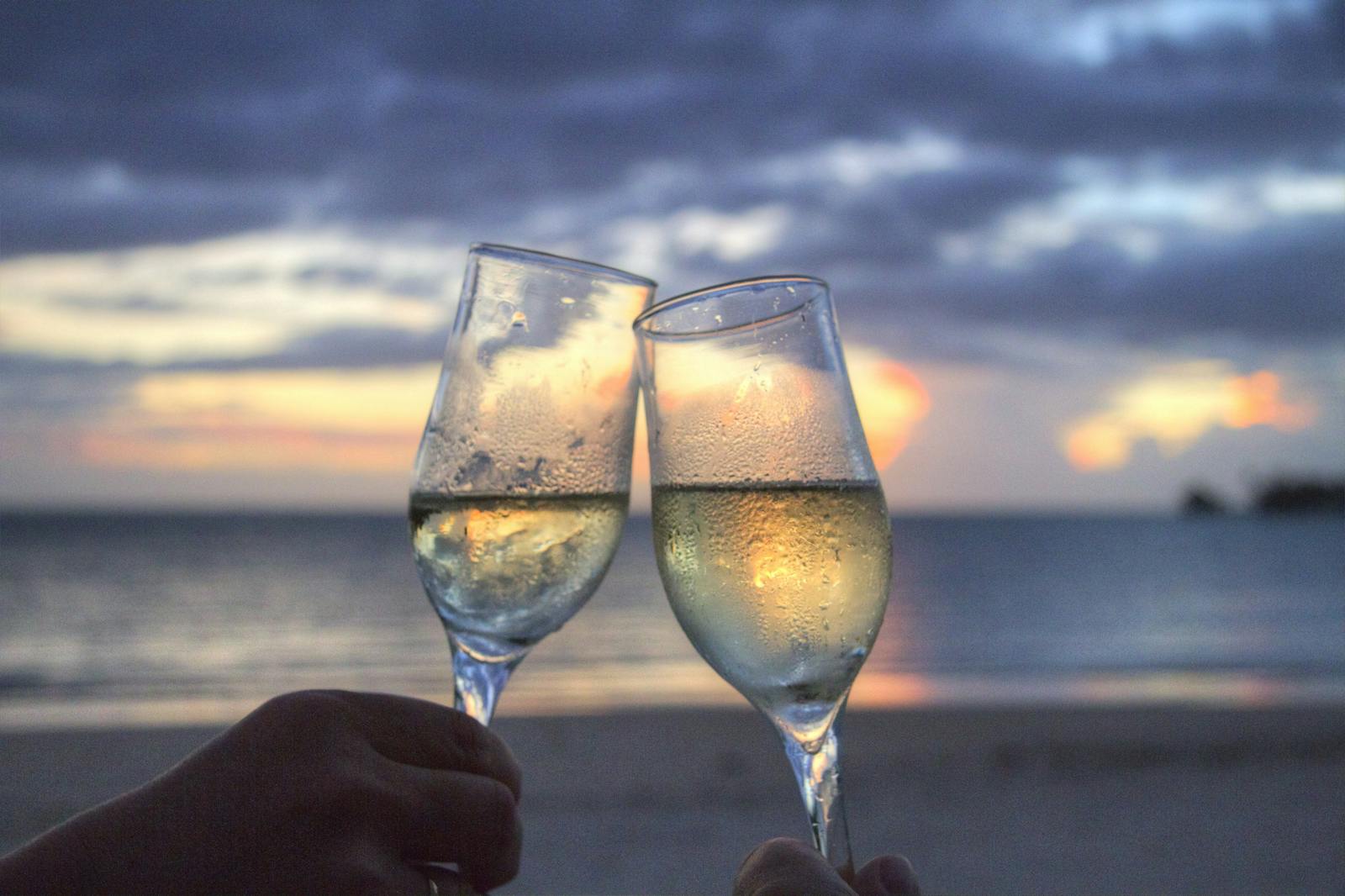 Two champagne glasses clink at sunset on a serene beach, evoking romance and celebration.